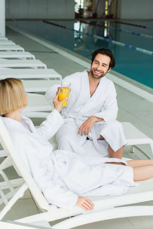 Smiling man clinking orange juice with girlfriend on deck chair in spa centerの写真素材