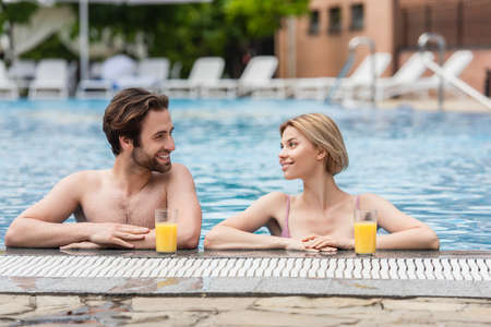 Cheerful woman in swimming pool looking at boyfriend near orange juice on poolside