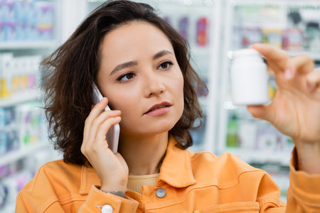 brunette woman holding bottle with medication while talking on smartphone in drugstoreの写真素材