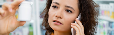 brunette woman holding bottle with medication while talking on smartphone in drugstore, bannerの写真素材