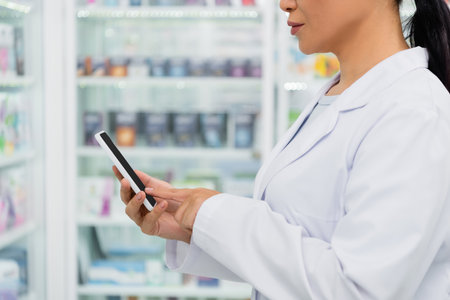 cropped view of pharmacist in white coat pointing at cellphone in drugstoreの写真素材