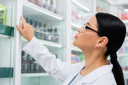 side view of asian pharmacist in glasses looking at medication on blurred shelvesの写真素材