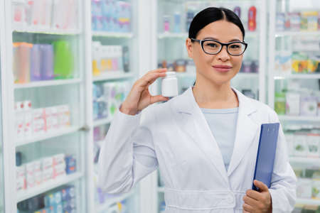 asian pharmacist in glasses smiling while holding bottle with medication and clipboardの写真素材