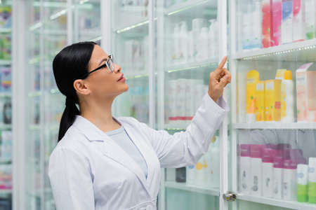brunette asian pharmacist in glasses pointing with finger at blurred shelves with medicationの写真素材