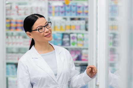cheerful asian pharmacist in glasses looking at blurred shelves with drugsの写真素材