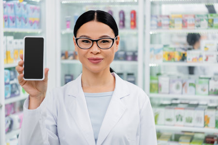 asian pharmacist in glasses smiling while holding cellphone with blank screen in drugstoreの写真素材
