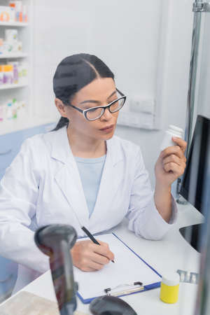 asian pharmacist in glasses holding bottle with medication while writing on clipboard in drugstoreの写真素材