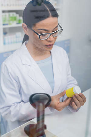 asian pharmacist in glasses and white coat looking at bottle with medication near scanner device at counterの写真素材