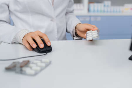 cropped view of pharmacist holding blister pack and using computer mouse in drugstore