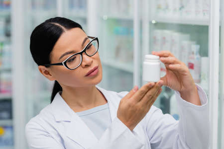 brunette asian pharmacist in glasses holding bottle with medicationの写真素材