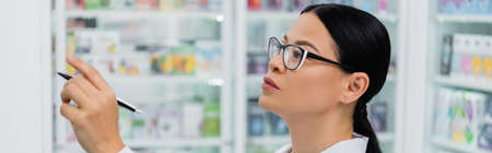asian pharmacist in glasses holding pen while pointing at drugs, bannerの写真素材