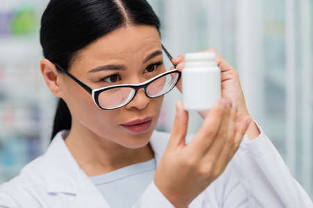 brunette asian pharmacist adjusting glasses and looking at bottle with medicationの写真素材
