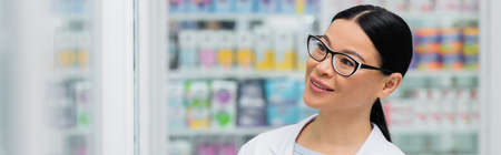 smiling asian pharmacist in glasses looking away in drugstore, bannerの写真素材