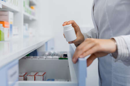 cropped view of pharmacist in white coat holding bottle with medication while opening drawer in drugstoreの写真素材