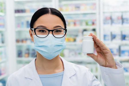 asian pharmacist in glasses and medical mask holding bottle with pillsの写真素材