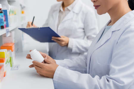 partial view of pharmacist in white coat holding bottle near blurred colleague writing on clipboard in drugstoreの写真素材