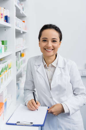 smiling pharmacist in white coat writing on clipboardの写真素材