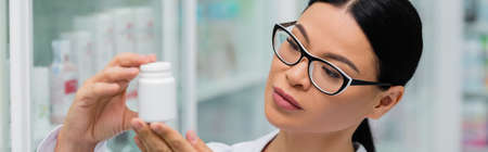 brunette asian pharmacist in glasses holding bottle with medication, bannerの写真素材