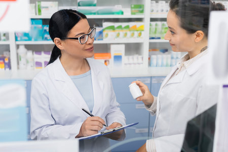 tattooed pharmacist in white coat holding bottle near smiling asian colleague in glasses writing on clipboard in drugstoreの写真素材
