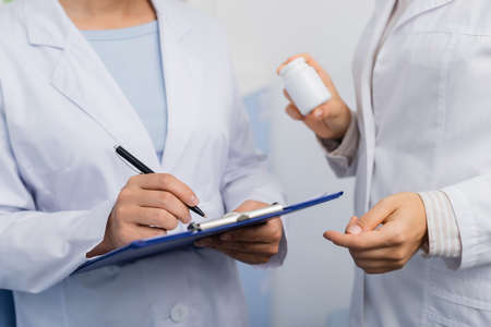 cropped view of pharmacist in white coat holding bottle near colleague writing on clipboard in...