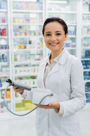 cheerful pharmacist in white coat holding digital tonometer in drugstoreの写真素材