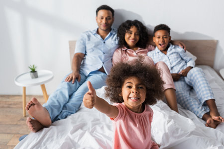 cheerful african american girl showing thumb up at camera near blurred family in bedroomの写真素材
