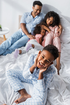 cheerful african american kid smiling at camera near blurred family in bedの写真素材