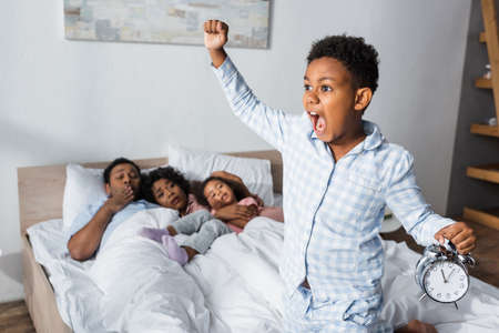 excited african american boy with alarm clock screaming and showing yeah gesture bedroomの写真素材