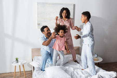 excited african american family in pajamas holding hands while fooling around on bed