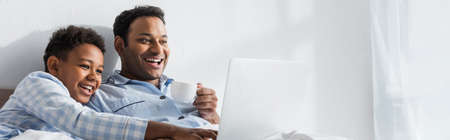 laughing african american boy pointing at laptop while watching movie with dad in bedroom, banner