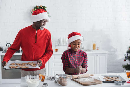happy african american man holding baking sheet with christmas cookies near grandson