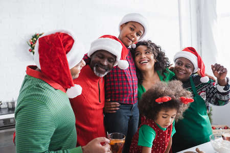 cheerful african american family in santa hats embracing and smiling in kitchen