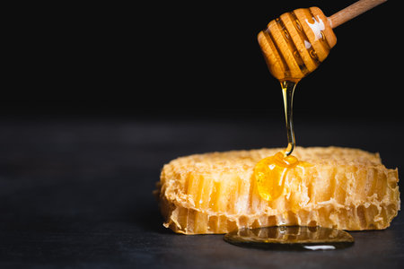 close up view of golden honey dripping on honeycomb from wooden dipper isolated on blackの写真素材
