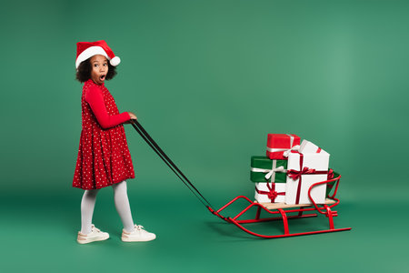 Amazed african american girl in santa hat pulling sleigh with gifts on green backgroundの写真素材