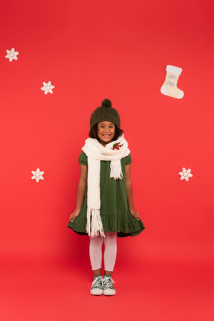 Cheerful african american kid in hat and scarf with mistletoe standing near decor on red backgroundの写真素材