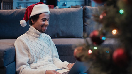 cheerful african american man in santa hat using laptop near blurred christmas tree