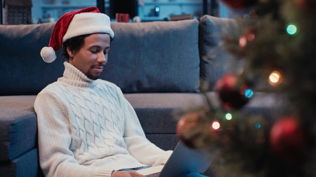 african american man in santa hat using laptop near blurred christmas tree