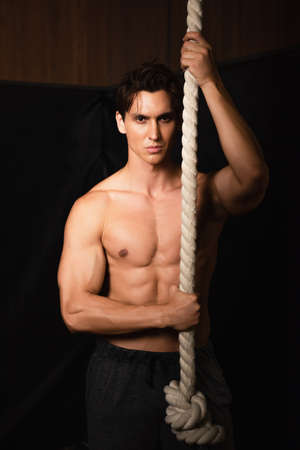 shirtless sportive man looking at camera while standing near gymnastic rope in gym