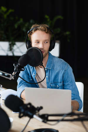 man in headphones talking near laptop and microphone in podcast studio