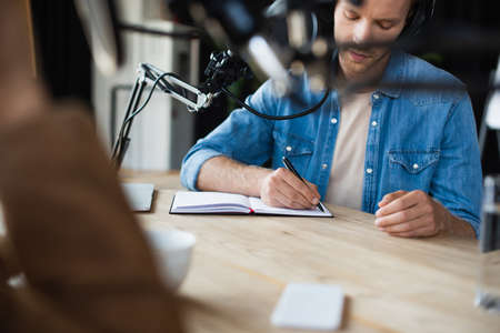 radio host in wireless headphones writing in notebook near blurred colleagueの写真素材