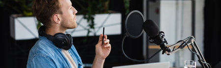 pensive influencer in headphones holding pen near microphone in studio, banner