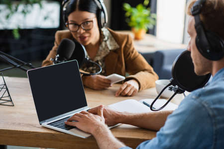 blurred radio host using laptop near asian colleague recording podcast in studioの写真素材