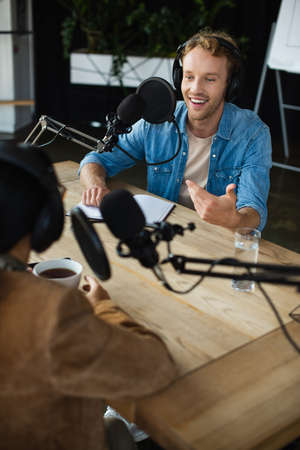 cheerful radio host in headphones looking at blurred colleague and gesturing during podcastの写真素材