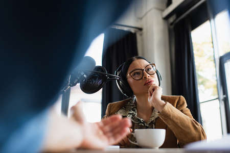 blurred radio host gesturing near asian colleague with cup of coffee