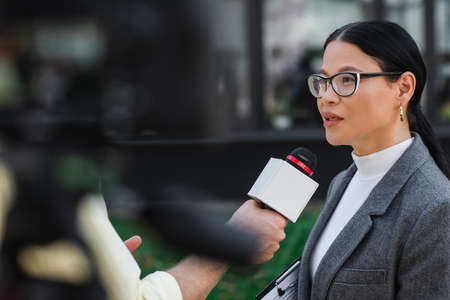 reporter holding microphone and taking interview of asian businesswoman in glasses outsideの写真素材