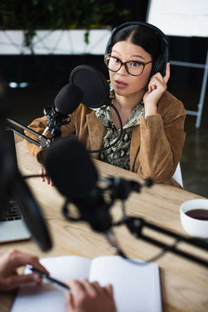 surprised asian radio host in glasses and headphones talking and looking at blurred colleague