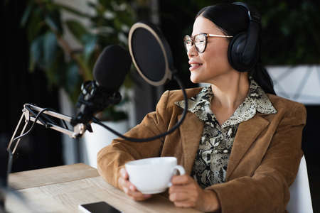 pleased asian radio host in glasses and headphones holding cup of coffee during podcastの写真素材