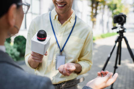 cheerful reporter holding microphone near blurred businesswoman gesturing while giving interviewの写真素材