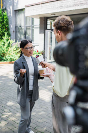 reporter holding microphone near cheerful asian businesswoman with paper cup near blurred digital cameraの写真素材