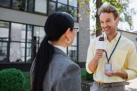 happy journalist smiling while holding microphone and looking at blurred asian businesswoman